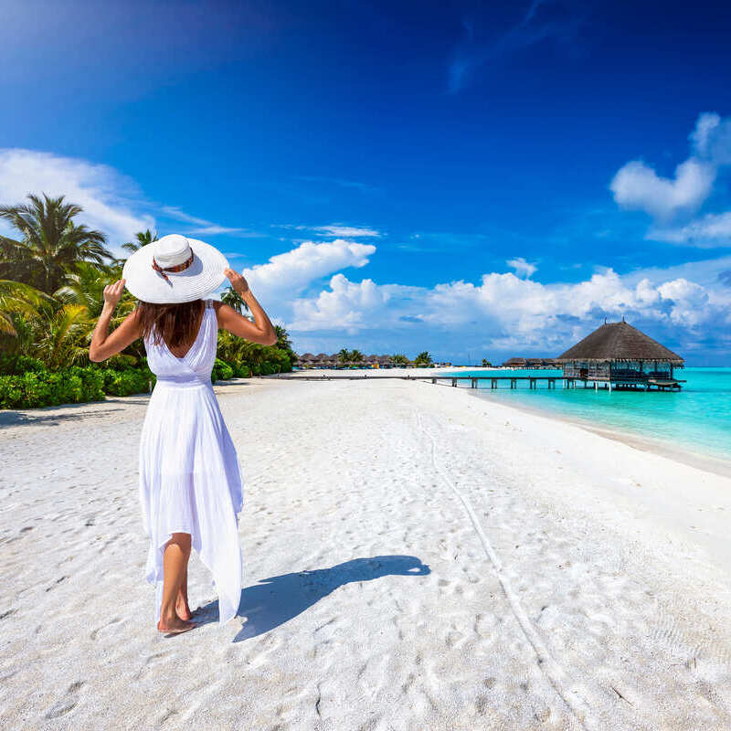 Młoda kobieta ubrana w białą sukienkę spacerującą po tropikalnej plaży w nieokreślonym tropikalnym miejscu, prawdopodobnie na Karaibach