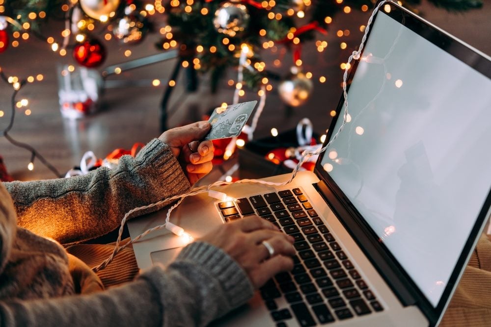 Świąteczny klient siedzi przy laptopie z kartą kredytową w dłoni i świątecznymi lampkami widocznymi w tle, opowiadając o sezonie świątecznych zakupów.