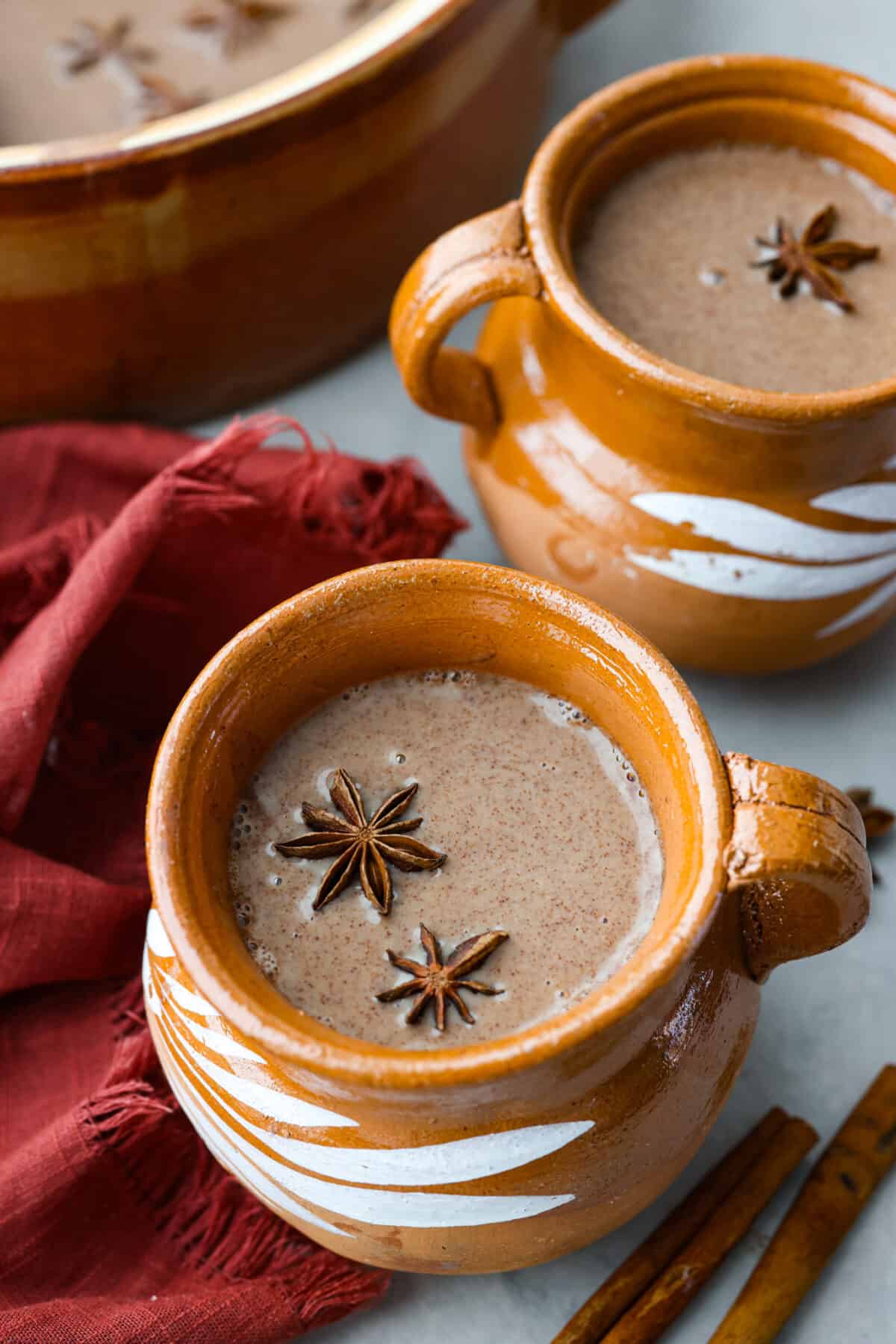 Champurrado w brązowym kubku.  Jest wypełniony anyżem i otoczony laskami cynamonu.