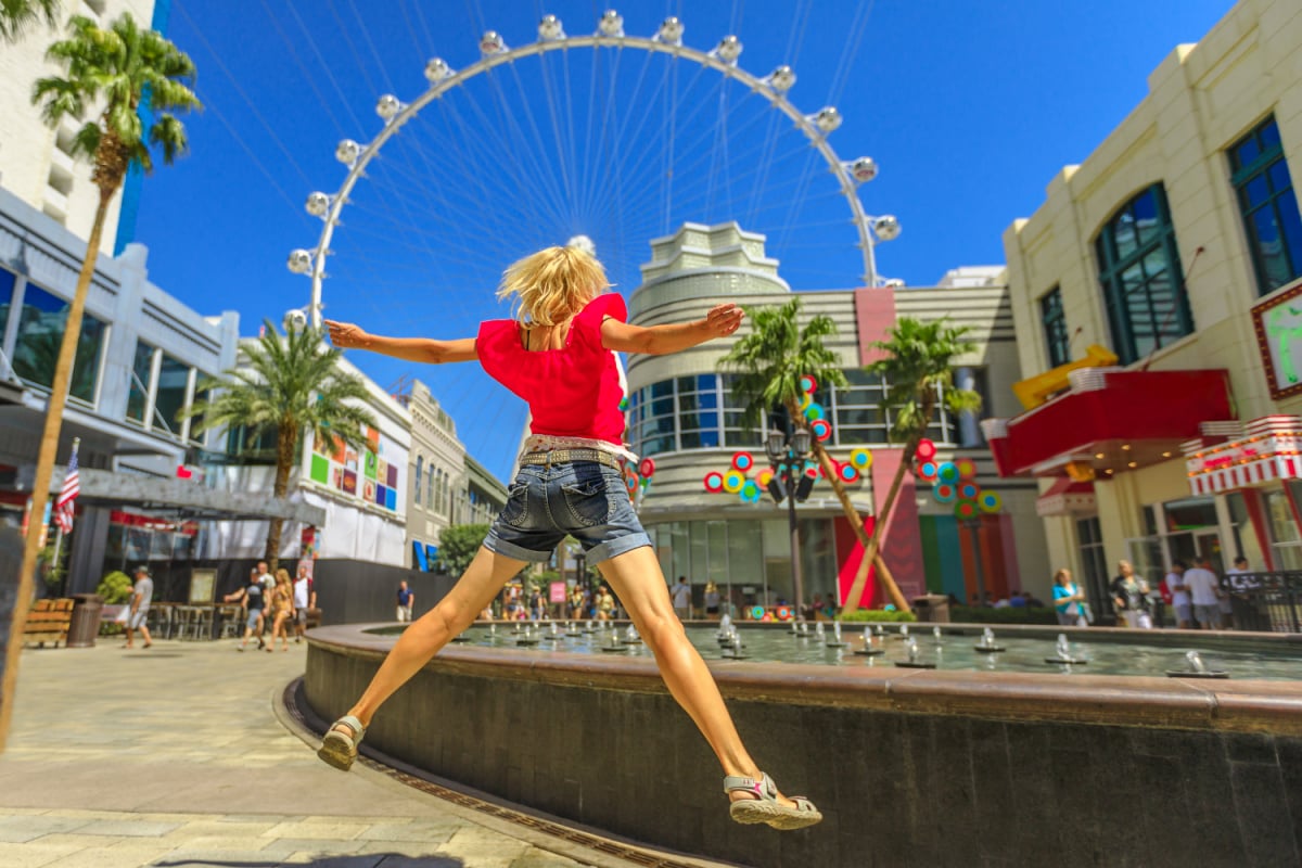 Blondynka skacząca w centrum Las Vegas w słoneczny dzień, błękitne niebo.  Diabelski młyn, największe na świecie koło obserwacyjne na rozmytym tle.  Szczęśliwy turysta w pejzażu miejskim Las Vegas, Nevada, USA