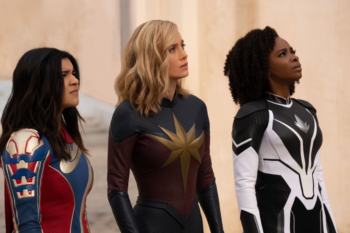 Iman Vellani jako Pani Marvel/Kamala Khan, Brie Larson jako Kapitan Marvel/Carol Danvers i Teyonah Parris jako Kapitan Monica Rambeau stoją razem w kostiumach, wszyscy patrzą w górę w filmie Marvel Cinematic Universe Marvels