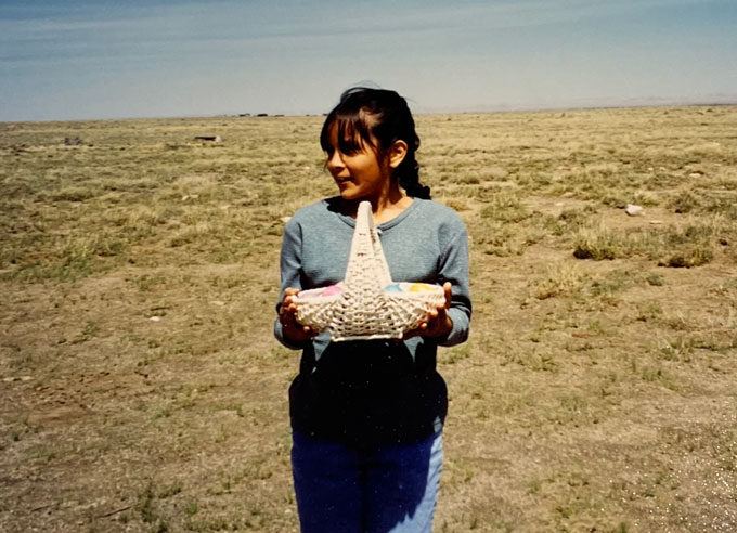 Zdjęcie Krystal Tsosie jako dziecka stojącego na polu w rezerwacie Navajo w Leupp w Arizonie, trzymającego biały kosz z plastikowymi pisankami.