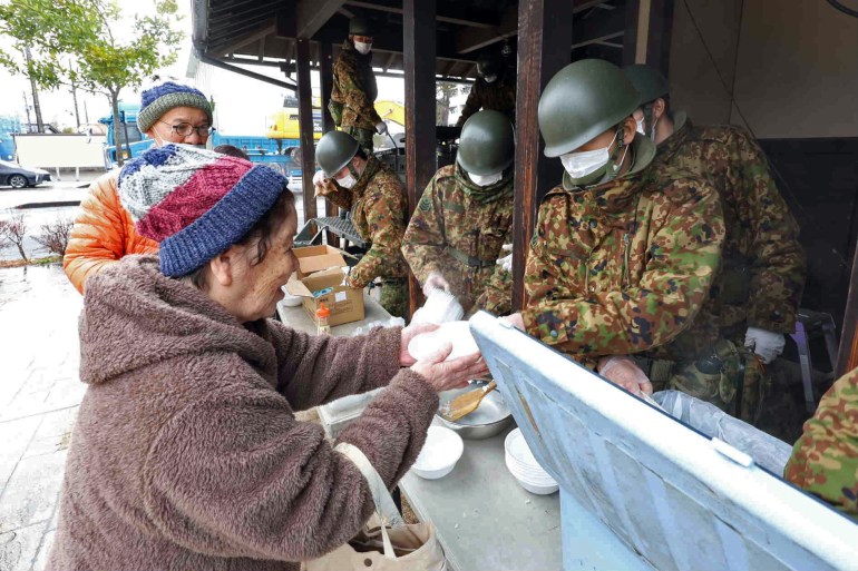 Japońscy żołnierze przygotowują w kuchni na świeżym powietrzu gorące posiłki dla osób, które przeżyły trzęsienie ziemi.  Starsza kobieta w płaszczu i wełnianej czapce zabiera część jedzenia.