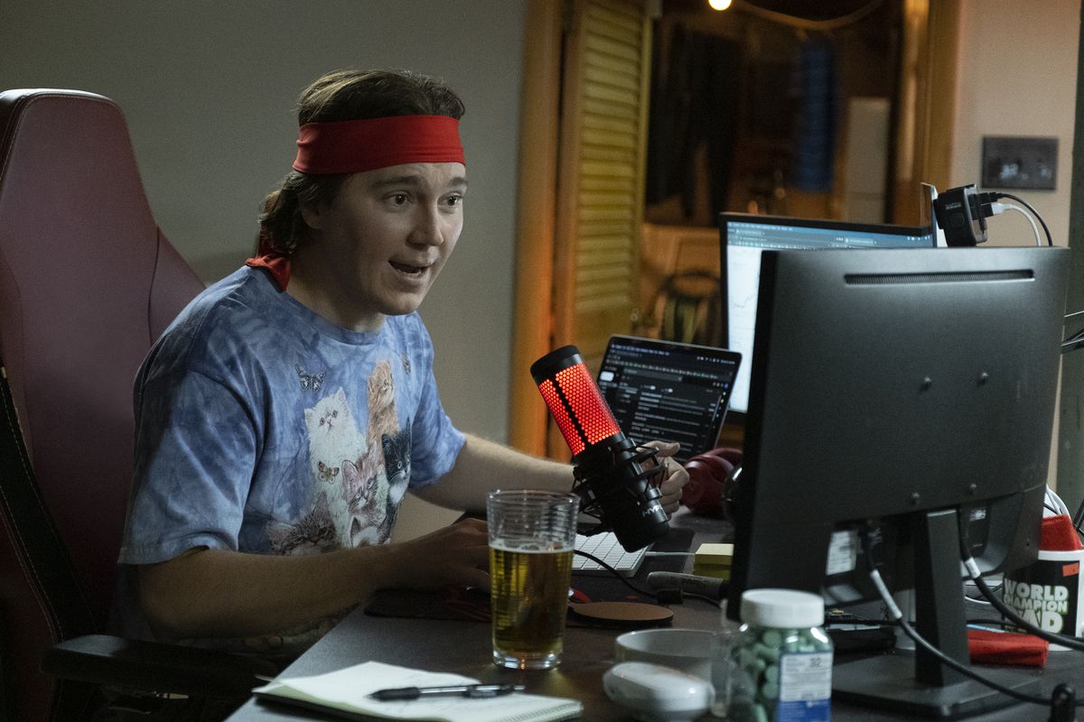 Paul Dano jako Keith Gill, znany również jako Roaring Kitty, siedzący przy biurku przy szeregu monitorów ze świecącym mikrofonem przed sobą, ubrany w nowatorską koszulkę z puszystymi kotkami i czerwoną bandanę zawiązaną wokół głowy w Dumb Money