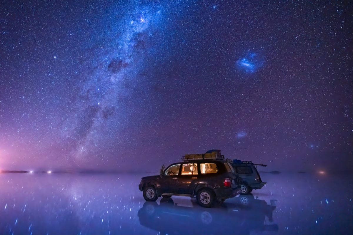 droga mleczna i gwiaździste niebo nad solniskiem Uyuni w Boliwii, z 2 SUV-ami zaparkowanymi na odblaskowej powierzchni jeziora.