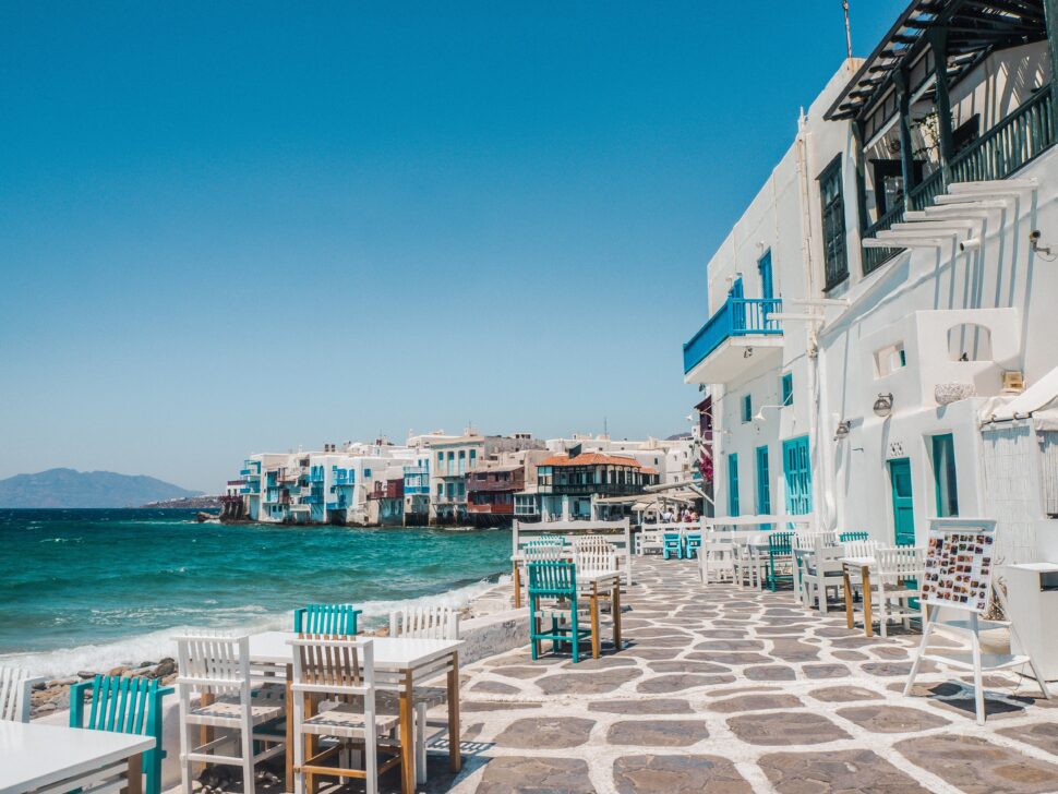 Słoneczne wybrzeże Mykonos z kolorowymi białymi i niebieskimi budynkami, kamiennym chodnikiem i krzesłami restauracyjnymi nad wodą