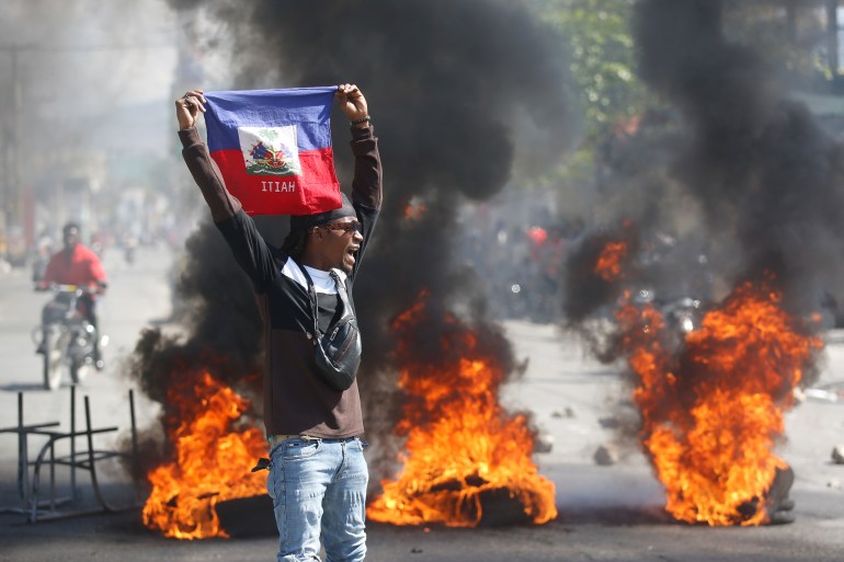 Mężczyzna trzymający nad głową flagę Haiti podczas zamieszek w Port-au-Prince.  Za nim płoną ognie i gęsty czarny dym