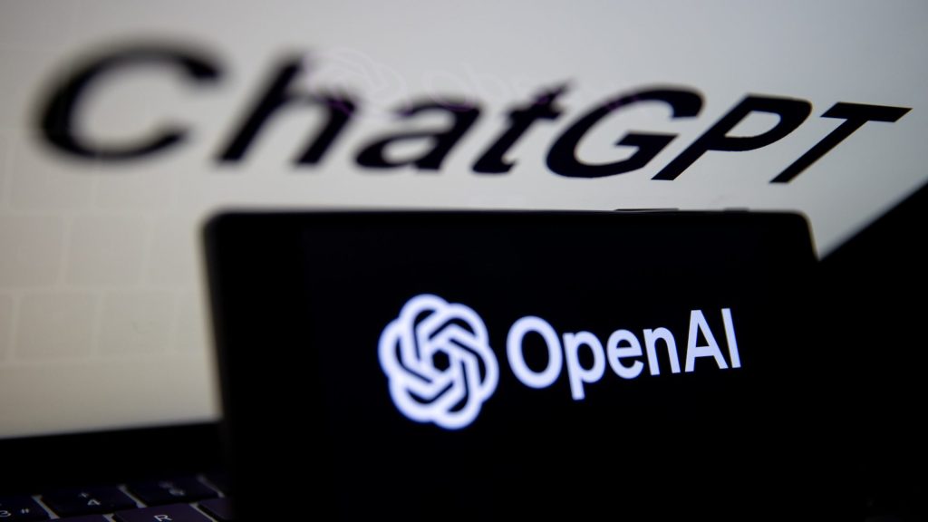 Logo OpenAI jest wyświetlane na ekranie telefonu komórkowego przed ekranem komputera z logo ChatGPT