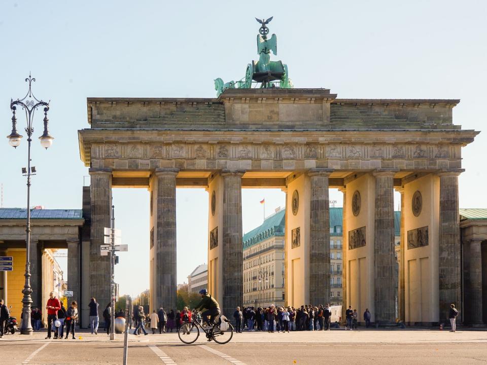 Światło słoneczne zagląda przez Bramę Brandenburską w Berlinie, a ludzie spacerują i jeżdżą na rowerach po ulicy przed nią