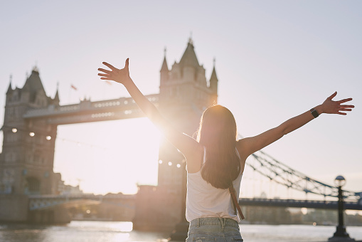 Ujęcie z tyłu przedstawiające kobietę stojącą obok Tower Bridge w Londynie o zachodzie słońca i wiwatującą