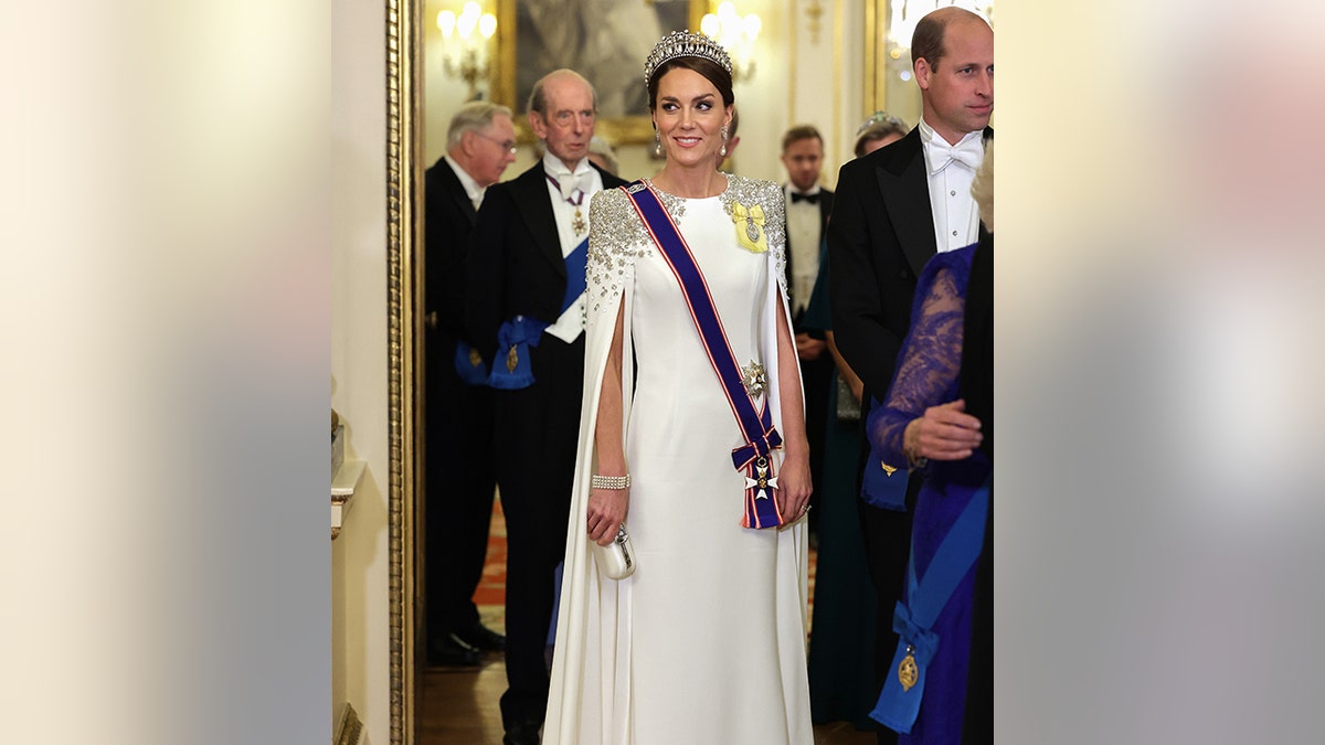 Księżniczka Katarzyna uśmiecha się, ubrana w długą białą suknię z klejnotami na ramionach i niebieską szarfą z księciem Williamem w tle