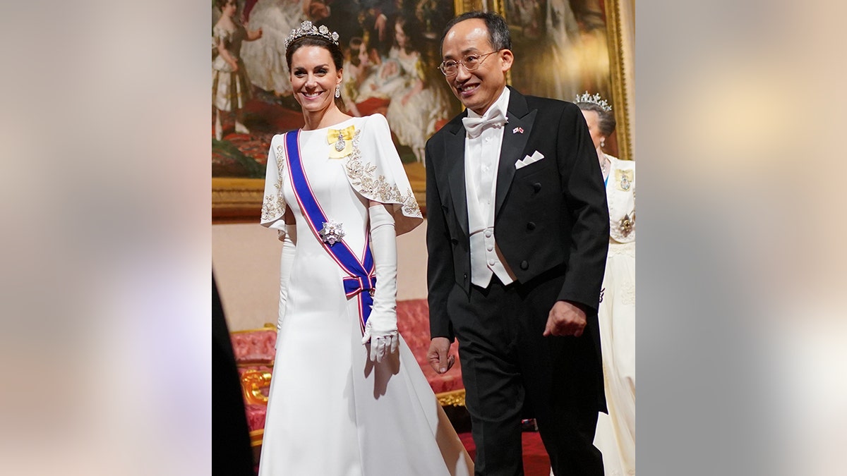 Księżniczka Katarzyna spaceruje w długiej białej sukni z niebieską szarfą i żółtą broszką obok mężczyzny w czarnym garniturze