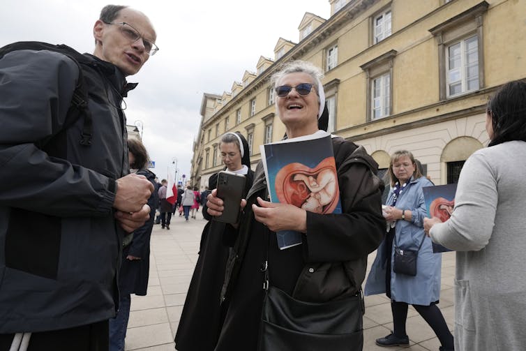 Kobieta w okularach przeciwsłonecznych i stroju zakonnicy trzyma zdjęcie dziecka w macicy, gdy rozmawia z mężczyzną na ulicy.