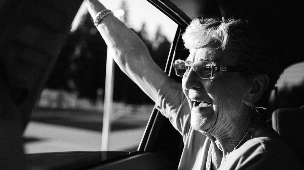 czarno-białe zdjęcie uśmiechniętej starszej kobiety w okularach machającej przez okno samochodu