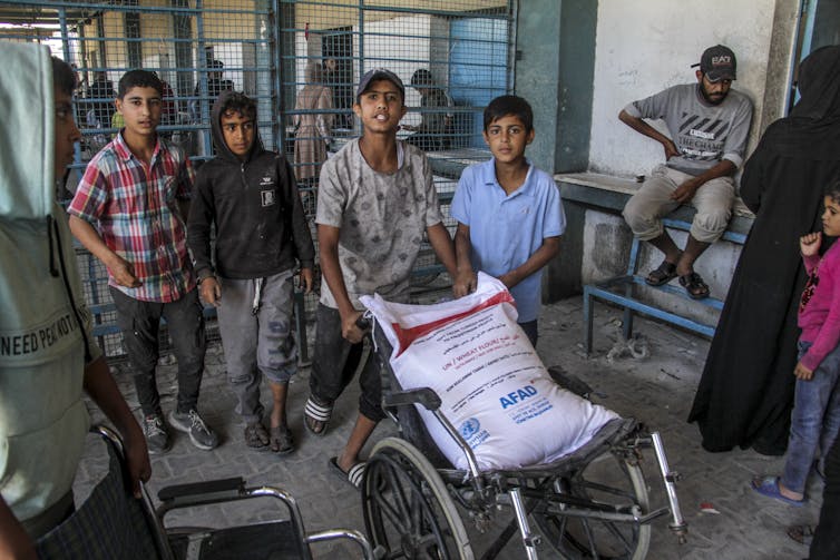 Czterech chłopców wyglądających na nastolatków patrzy w kamerę i pcha wózek inwalidzki z dużą białą torbą.