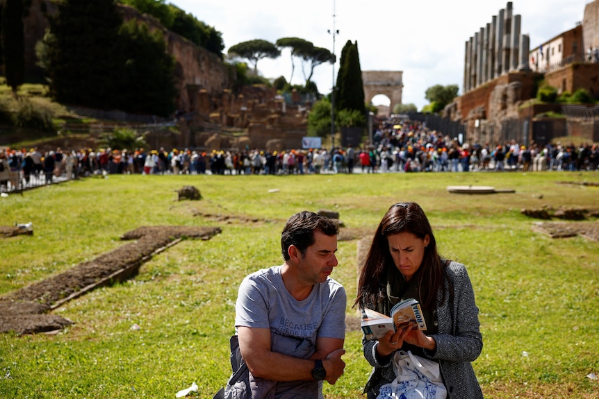 Turyści czytają przewodnik turystyczny po Rzymie, siedząc w pobliżu tłumów w Koloseum w Rzymie.