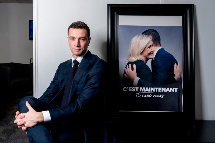 Jordan Bardella przed fotografią przedstawiającą go i Marine Le Pen przytulających się