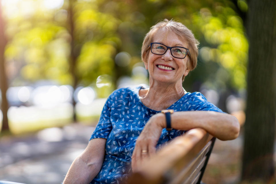 Starsza kobieta w okularach i wzorzystej bluzce siedzi na ławce w parku i uśmiecha się do kamery.  W tle widoczne są drzewa i światło słoneczne