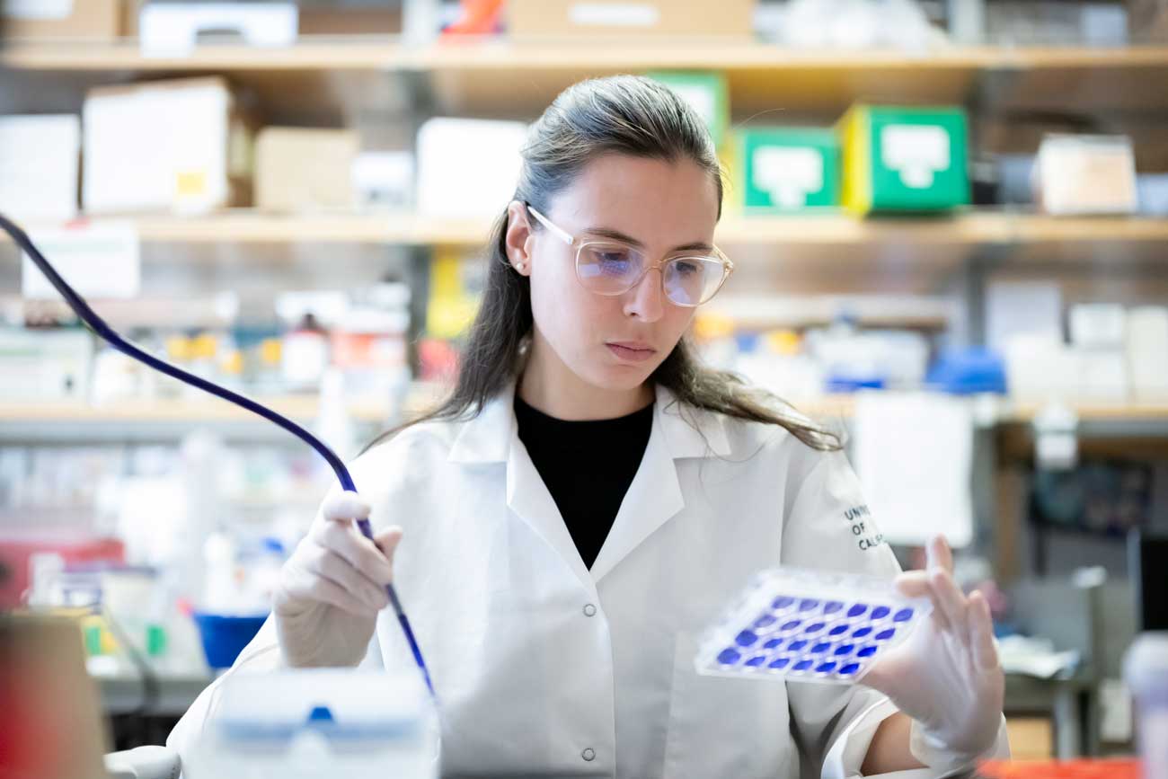 Jovanka Gencel-Augusto ma na sobie fartuch laboratoryjny i okulary ochronne, gdy zarządza sprzętem laboratoryjnym i próbkami.