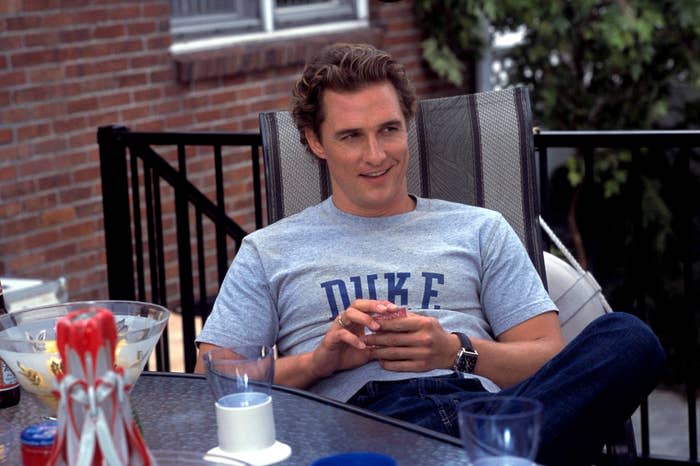 Matthew McConaughey siedzi na krześle na patio, ubrany w "Książę" T-shirt i dżinsy, trzymając drinka i uśmiechając się