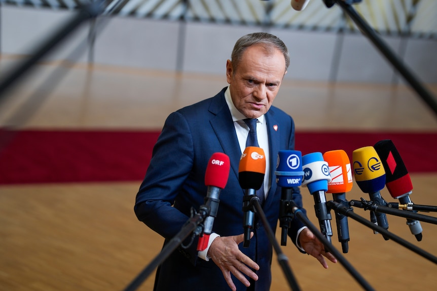 Premier Polski Donald Tusk stoi przed siedmioma różnymi mikrofonami, ubrany w granatowy garnitur i krawat.