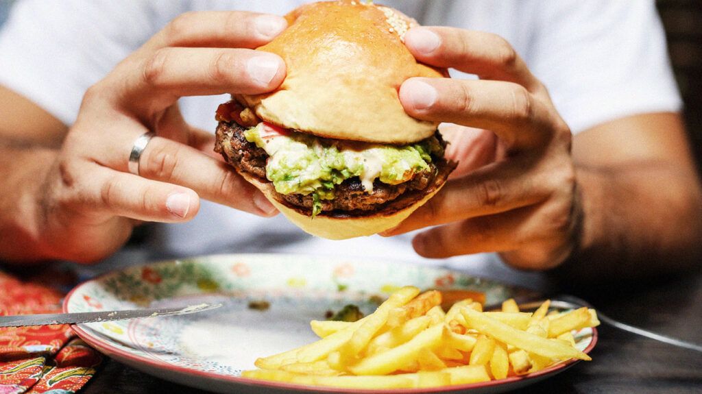 Osoba trzymająca cheeseburgera nad talerzem frytek, oba przedstawiające potrawy o wysokiej zawartości tłuszczu