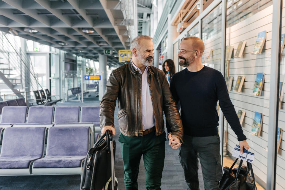 Para gejów spacerująca z bagażem, trzymająca się za ręce i patrząca na siebie, omawiająca plany wakacyjne, gdy razem przechodzą przez terminal lotniska i przygotowują się do złapania lotu.