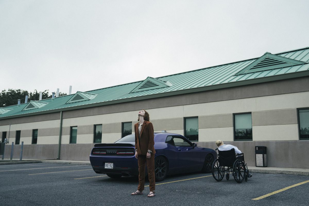 Emma Stone stoi z odchyloną do tyłu głową obok fioletowego Dodge’a Challengera przed anonimowym budynkiem medycznym.  Jest postać na wózku inwalidzkim, odwrócona tyłem do kamery