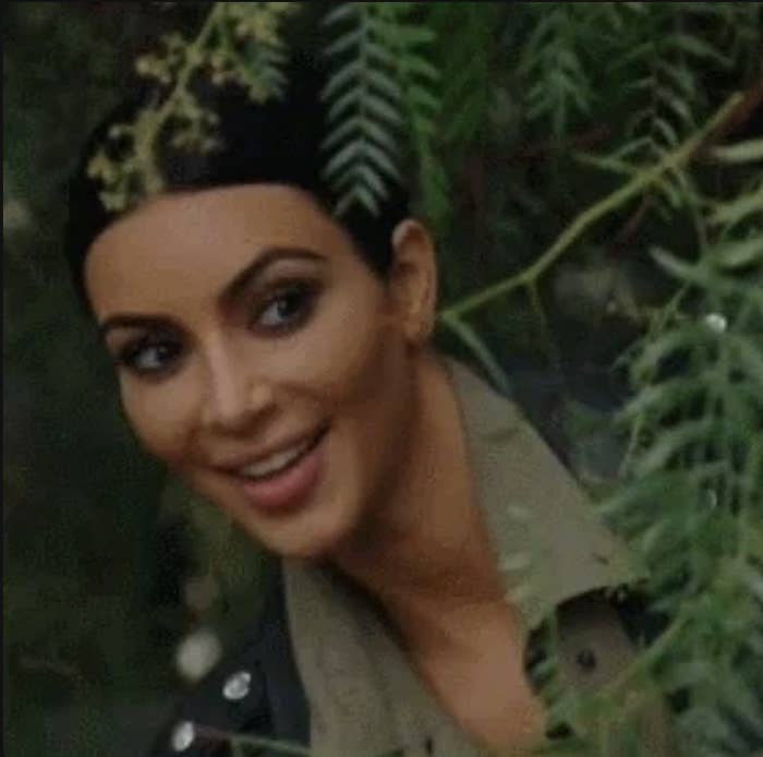 Kim Kardashian spogląda przez gałęzie drzew i uśmiecha się.  Nosi marynarkę, a pod nią koszulę z klapami