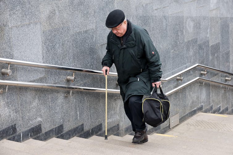 Starszy mężczyzna wspinający się po wielu schodach, trzymający laskę.