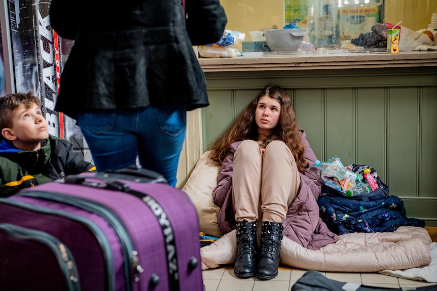Nastolatka siedzi na podłodze oparta o ladę, obok walizka i młody chłopak