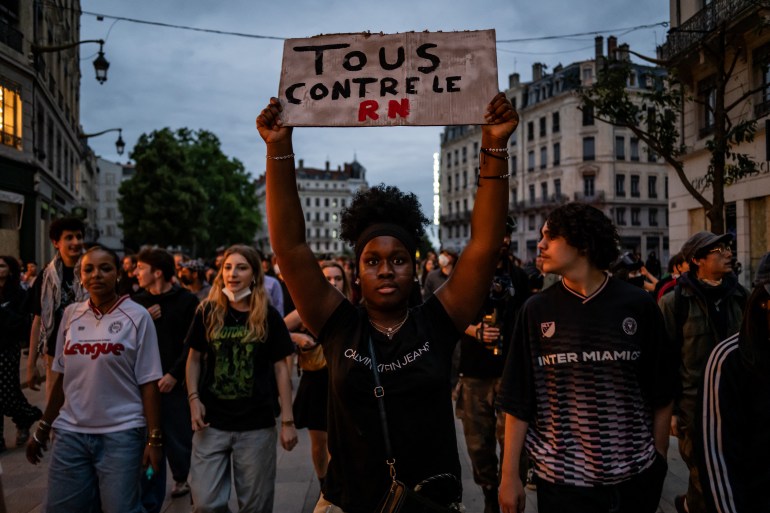 Protestujący w Lyonie trzyma plakat z napisem „Wszyscy przeciwko RN”.  Za nią idą ludzie.