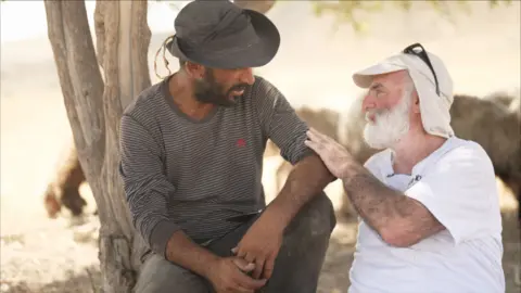 Ahmad, palestyński rolnik ubrany w koszulkę z długim rękawem i kapelusz, siedzi z Gilem i mężczyzną z białą brodą. Gil położył rękę na ramieniu Ahmeda i rozmawiają, siedząc pod drzewem.