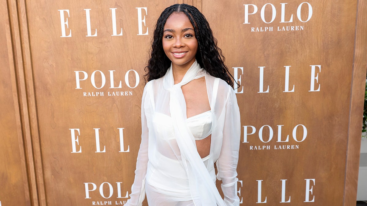 Skai Jackson w białym biustonoszu i prześwitującej białej bluzce uśmiecha się na dywanie