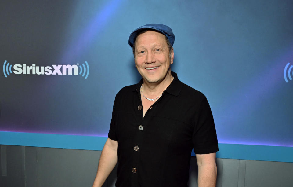 Rob Schneider w czarnej koszuli i niebieskiej czapce przed ścianą z logo SiriusXM