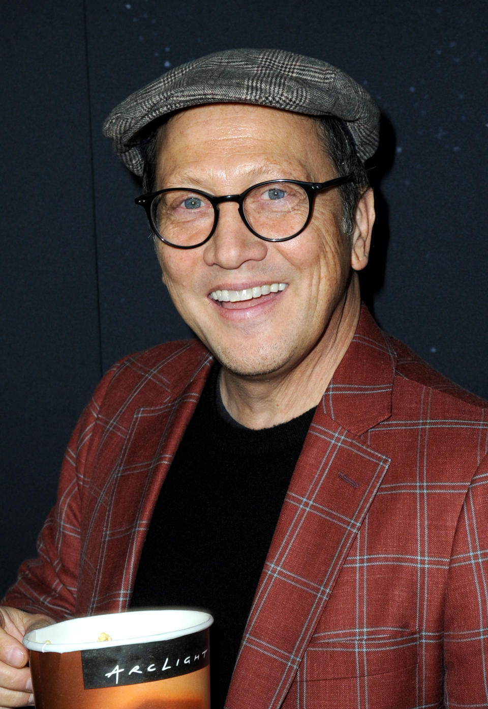 Rob Schneider uśmiecha się, trzymając kubek, ubrany w kraciastą marynarkę, czarną koszulę, okrągłe okulary i czapkę z daszkiem