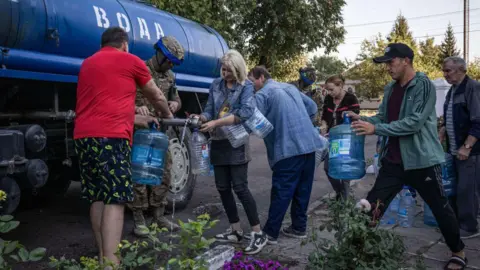 Getty Images Ludzie napełniają butelki wodą przed szkołą, która jest używana jako schron przeciwbombowy w kontrolowanym przez Ukrainę mieście Sudza, 18 sierpnia 2024 r.