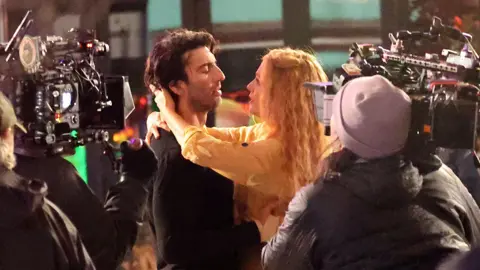 Getty Images Justin Baldoni i Blake Lively na planie It Ends With Us. Justin ma luźne brązowe włosy i nosi czarny sweter. Blake, która ma kręcone blond włosy, trzyma ręce na jego szyi i patrzy mu w oczy. Są otoczeni przez operatorów kamery, którzy ich filmują. 