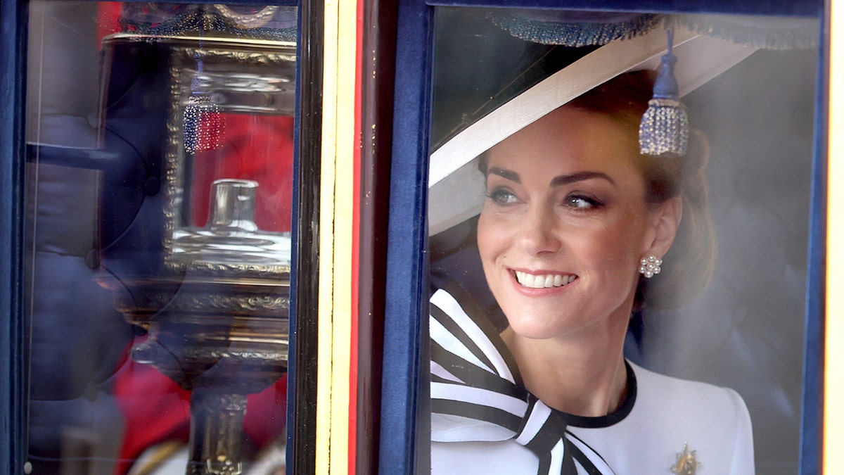 Kate Middleton w białej sukni z pasiastą kokardą spogląda i uśmiecha się do tłumu podczas przybycia Trooping the Colour