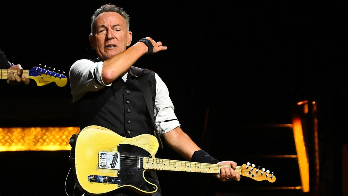 Bruce Springsteen występuje na scenie w Pittsburghu, ubrany w czarną kamizelkę i grający na żółtej gitarze, machając ręką w powietrzu