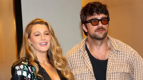 Getty Images Blake Lively i Brandon Sklenar na zdjęciu w Nowym Jorku w sierpniu. Blake to kobieta po trzydziestce z długimi, kręconymi blond włosami. Nosi pomalowaną skórzaną kurtkę i się uśmiecha. Brandon, 34-letni mężczyzna, ma kudłate brązowe włosy i krótki, zarośnięty zarost. Nosi brązowe okulary przeciwsłoneczne i brązową koszulę w kratkę gingham na czarnej kamizelce. 