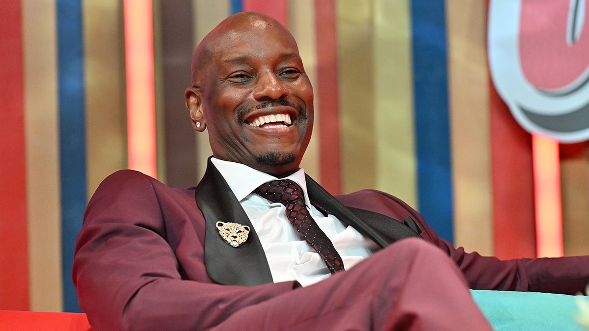 Tyrese Gibson uśmiecha się na scenie w bordowym garniturze