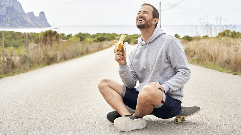 uśmiechnięty mężczyzna siedzi na zewnątrz na chodniku i je banana