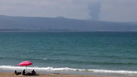 Reuters Rodzina siedzi na plaży w Tyrze w Libanie, gdy dym unosi się po libańskiej stronie granicy z Izraelem podczas intensywnej wymiany ognia między Hezbollahem a izraelskim wojskiem, 25 sierpnia 2024 r.