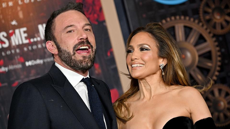 Ben Affleck w czarnym garniturze i krawacie trzyma usta otwarte na dywanie, podczas gdy Jennifer Lopez w czarnej sukience patrzy na niego z uwielbieniem